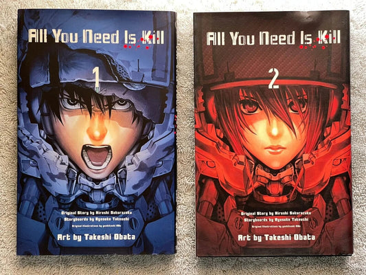 Two physical copies of the manga volume 'All You Need Is Kill' Volume 1 and Volume 2, displayed side-by-side on a textured beige carpet. The left copy (Volume 1) features a dark blue and black cover with a character in a helmet, while the right copy (Volume 2) features a dark red and black cover with a character in a red mechanical suit.